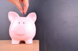 A coin is deposited into a pink piggybank with a blank blackboard in the background.